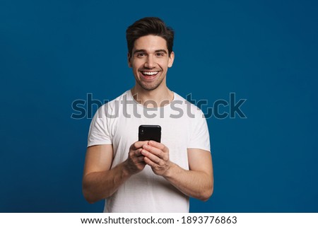 Happy unshaven guy smiling while using mobile phone isolated over blue background