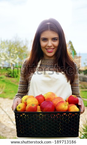 Portrait of a cheerful woman holding basket with apples in garden