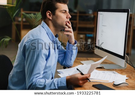 Pensive attractive young entrepreneur looking through paperwork while sitting at the desk