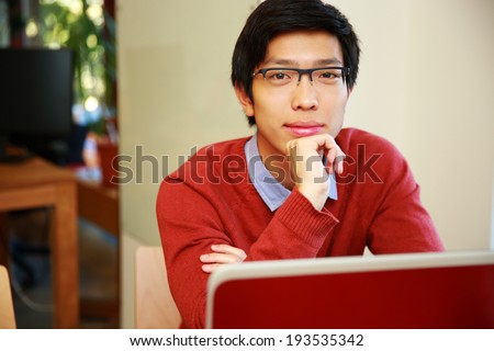 Handsome asian man sitting at the table with laptop and looking at camera