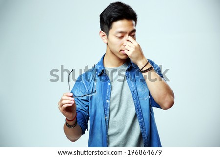 Asian man holding glasses and rubbing his eyes on gray background