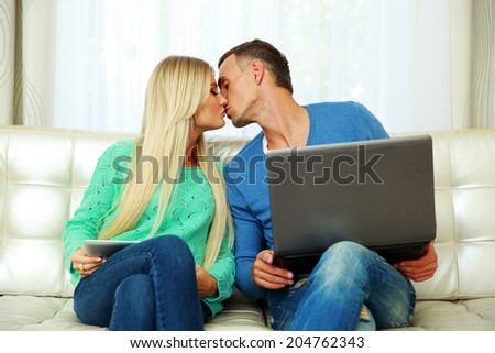 Young couple sitting at the sofa with laptop and tablet computer, and kissing
