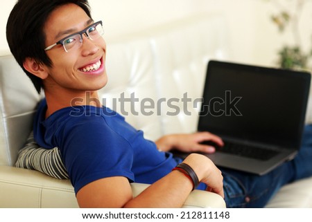 Cheerful asian man lying on the sofa with laptop at home