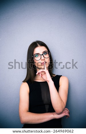 Portrait of a beautiful woman in glasses with finger over lips. Making shhh sign. Be quiet!!! Standing over gray background. Wearing black fashion dress. Looking at the camera.