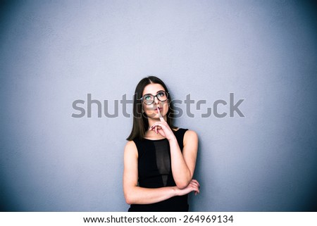 Young charming woman in glasses with finger over lips standing over gray background. Looking up at copyspace. Wearing in black fashion dress