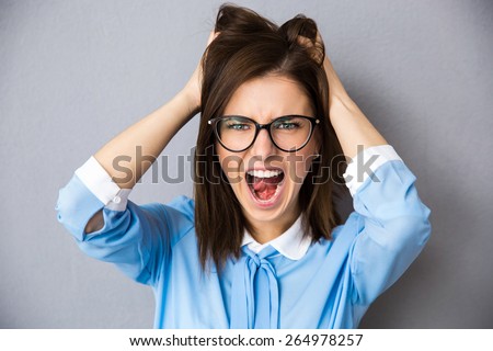 Angry businesswoman touching her hair and screaming over gray background. Wearing in blue shirt and glasses. Looking at camera