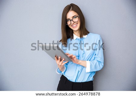 Smiling businesswoman standing with tablet computer over gray background. Wearing in blue shirt and glasses. Looking at camera