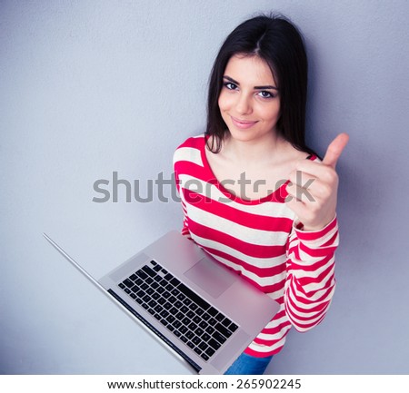Smiling woman standing with laptop and showing thumb up over gray background. Looking at the camera