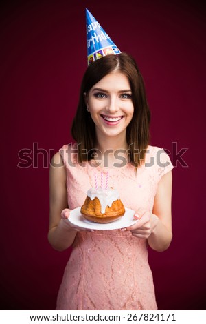 Smiling pretty woman holding cake with candles over pink background. Looking at camera. Celebrating her birthday! Wearing in pink dress