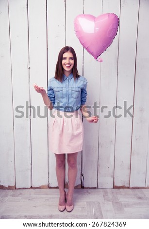 Full length portrait of a young smiling woman holding cookies and pink heart shaped balloon with wooden wall on background