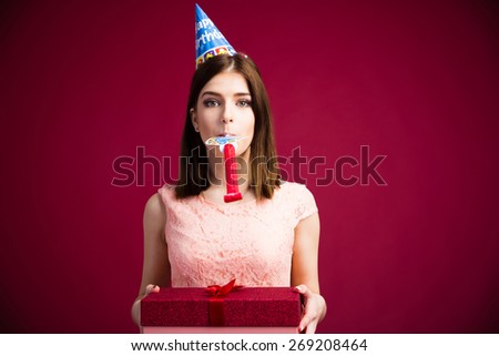 Woman blowing in whistle and holding gift box over pink background. Looking at camera. Wearing in dress