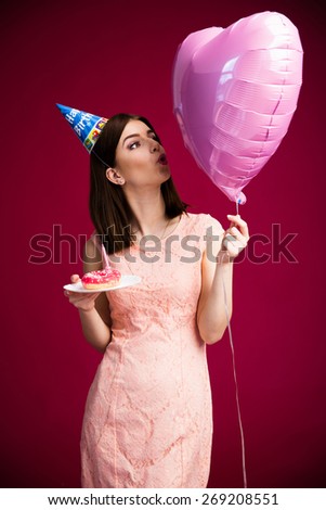 Woman holding donut with candle and heart shaped balloon over pink background. Wearing in dress. Blowing on balloon