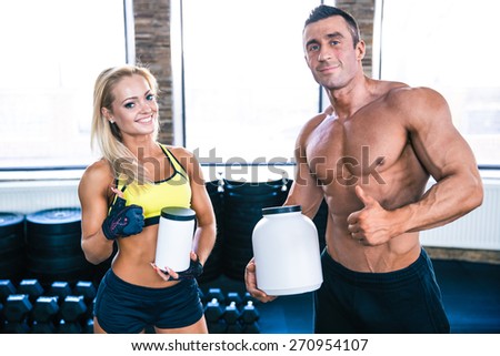 Man and woman holding container with sports nutrition and showing thumb up at gym