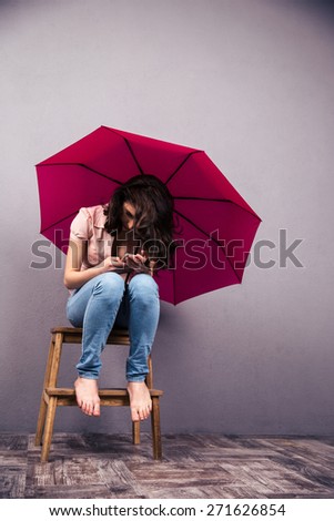 Woman sitting on the chair with pink umbrella and using smartphone at studio. Wearing in jeans and shirt. Barefoot