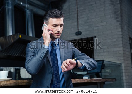 Confident businessman speaking on the phone and looking on the wristwatch in cafe