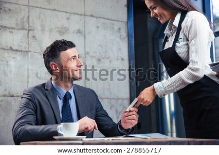 Cheerful confident man giving bank card to smiling female waiter in restaurant