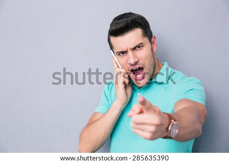 Casual young man talking on the phone and pointing finger at camera over gray background