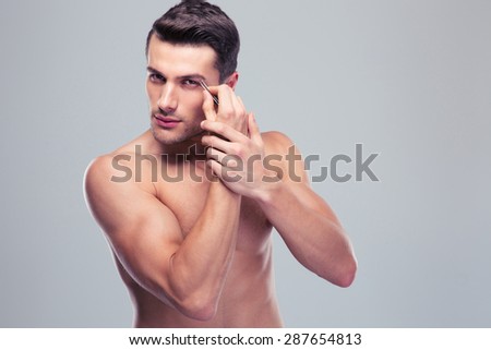 Handsome man removing eyebrow hairs with tweezing over gray background