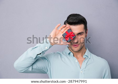 Young casual man covering his eye with gift box over gray background and looking at camera