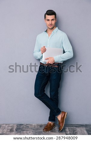 Full length portrait of a young handsome man standing and holding laptop over gray background