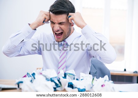 Angry businessman sitting at the table with crumpled paper and shouting in office