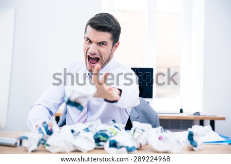 Angry businessman sitting at the table and throwing crumpled paper on camera