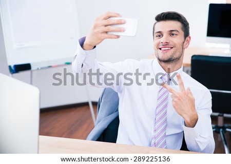 Happy businessman sitting at the table and making selfie photo on smartphone in office