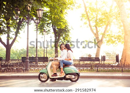 Sideview of a trendy couple riding a scooter in the morning. Sun is shining through trees