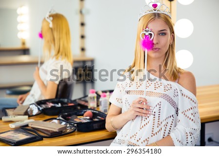 Attractive makeup artist wearing in queen crown covering her eye with magic wand in beauty salon. Looking at camera