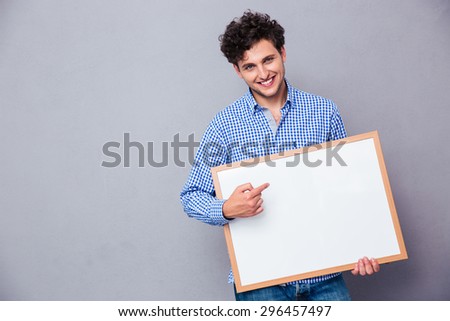 Cheerful man holding blank board and poiting finger on it over gray background and looking at camera