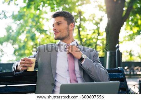 Portrait of a happy businessman sitting on the bench with laptop and drinking coffee outdoors. Looking away