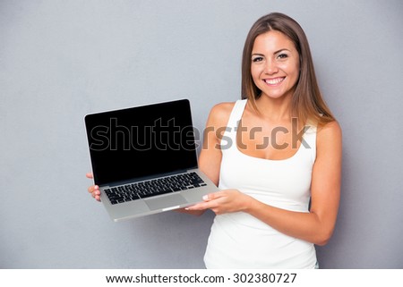 Happy young girl showing blank laptop screen over gray background. Looking at camera