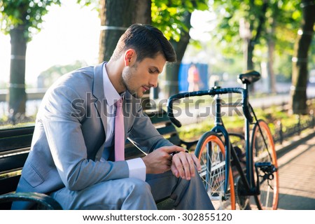Young businessman sitting on the bench outdoors and looking on wrist watch
