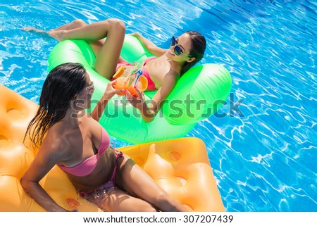 Portrait of a two girlfriends sunbathing on air mattress and drinking cocktails in swimming pool