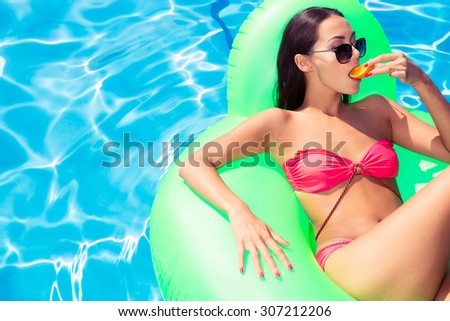 Portrait of attractive woman lying on air mattress and eating orange in swimming pool