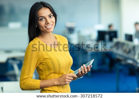Portrait of a cheerful casual businesswoman in sweater standing with tablet computer in office and looking at camera