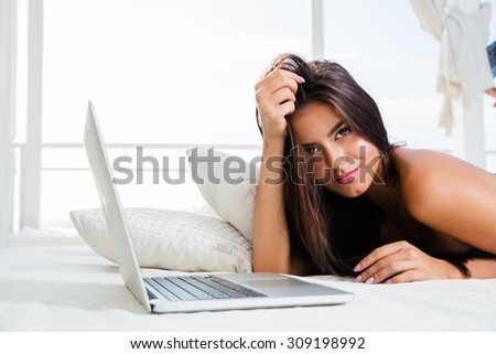 Portrait of a sexy young girl lying on the bed with laptop and looking at camera