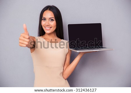Portrait of a cheerful woman holding laptop and showing thumb up over gray background. Looking at camera