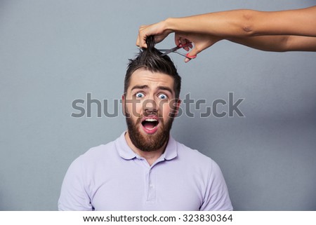 Portrait of a man looking scared while female hands cutting his hair over gray background