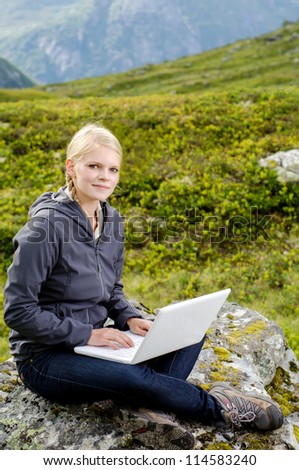 young blond woman sits with a laptop on a stone before mountain landscape