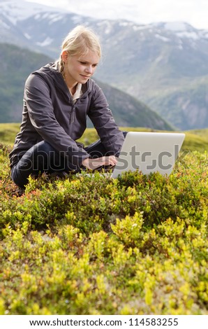 young blonde woman sits with laptop in alpine meadow before mountain landscape