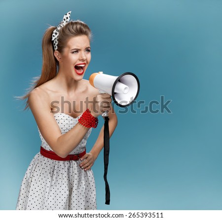 Wroth pin-up girl yells through megaphone, mouthpiece, speaking trumpet. Filmmaking or film production concept / photo set of young American pin-up model on blue background