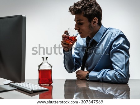 Young alcoholic man drinking whiskey sitting drunk at office with computer / photo of businessman addicted to alcohol at the workplace, depression and crisis concept