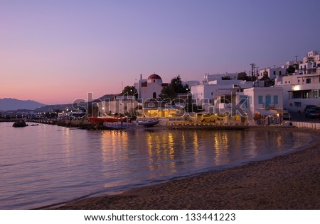 Sunset on the sea near the island of Mykonos in Greece and the night lights of restaurants and churches on the beach ...