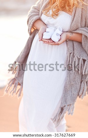 Beautiful pregnant woman with long blond hair standing on the beach at sunset in white dress and covering in wool grey pleid.