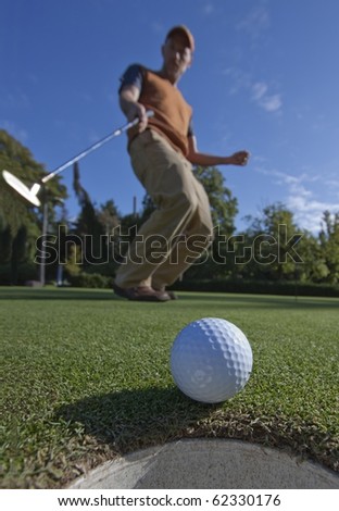 A golfball just as it\'s about to drop into the cup, golfer in the background arching as it drops