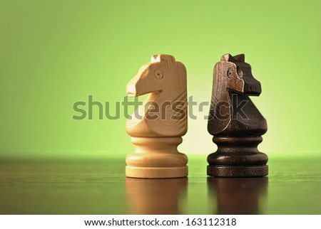 Two wooden knight chess pieces in opposing light and dark colours standing back to back on a reflective wooden surface against green with copyspace