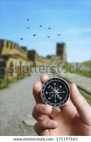 Conceptual image of the hand of a man using a compass to navigate and find the direction while sightseeing abroad with the ruins of an ancient castle in the background