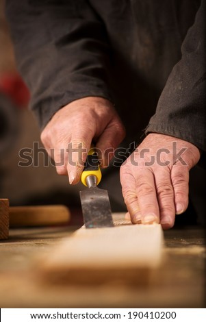 Carpenter using a chisel on a plank of wood to shape and cut a groove as he manufactures an article in his woodworking workshop, view of the hands and chisel