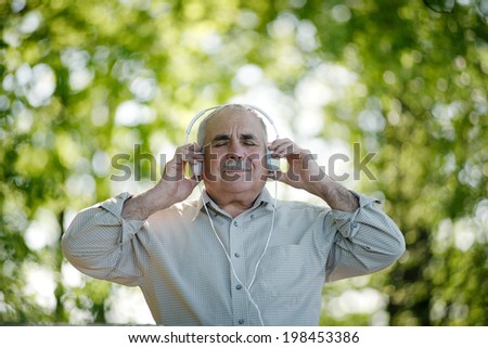 Senior man with a blissful expression stands stands outdoors in a wooded garden listening to music on his headphones smiling in satisfaction